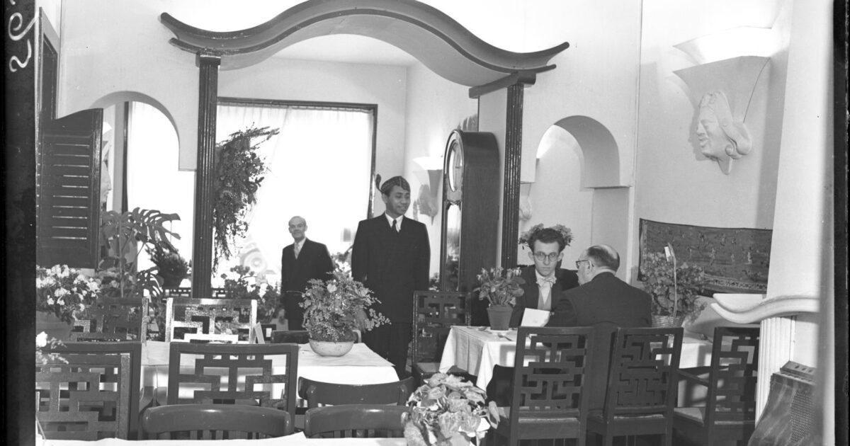 Black-and-white 1948 photograph of the dining room of an Indonesian restaurant in Amsterdam, with laid tables, lattice-backed chairs, an arched room divider and a waiter standing at the back