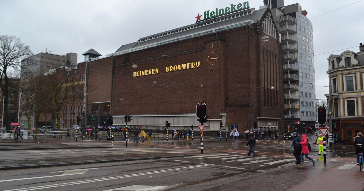 The brick Heineken Experience building in Amsterdam overlooks wet tram tracks, with pedestrians crossing beneath its rooftop sign.