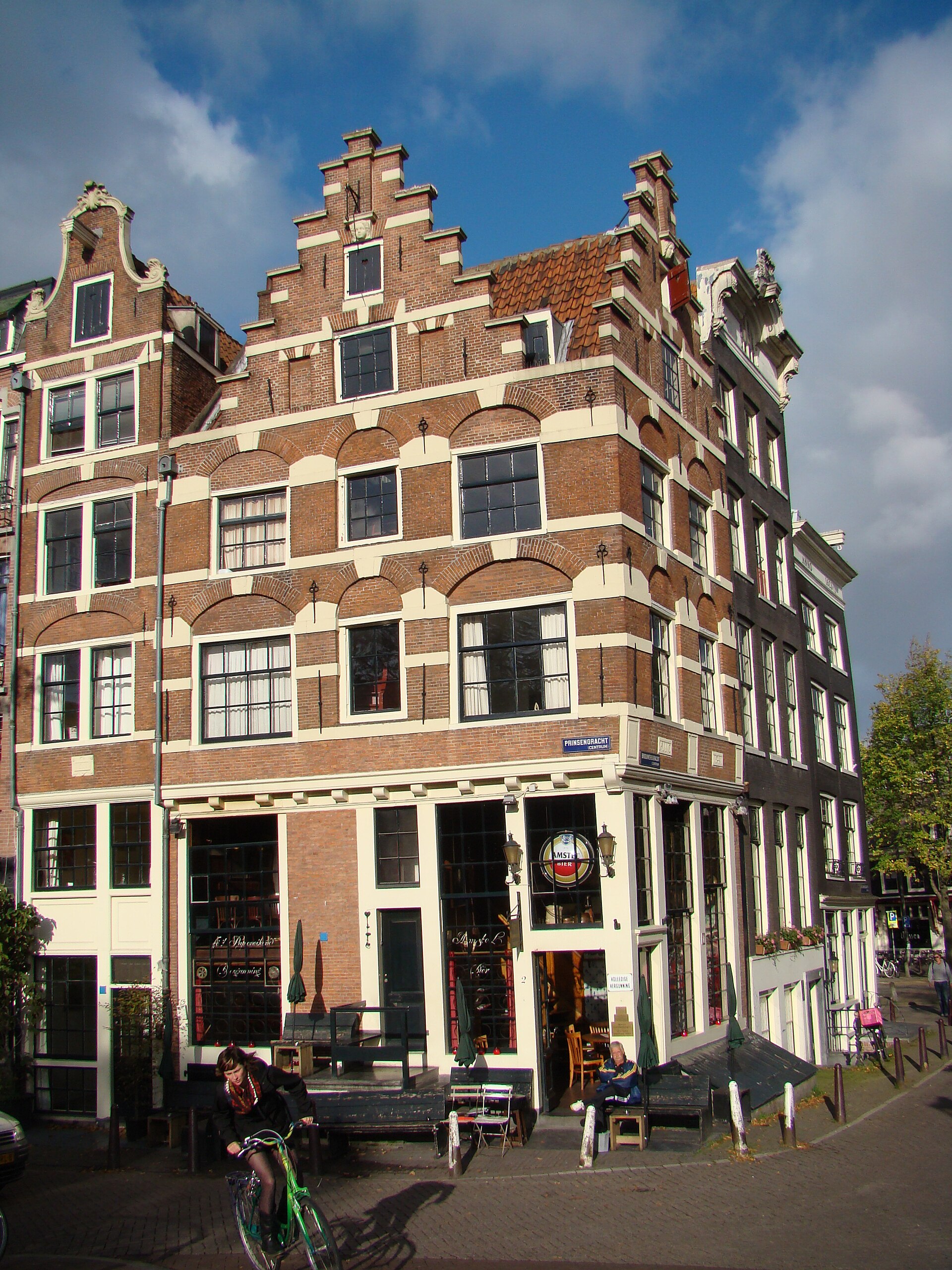 Cafe 't Papeneiland occupies a stepped-gable corner building in Amsterdam, with chairs outside and a cyclist passing.