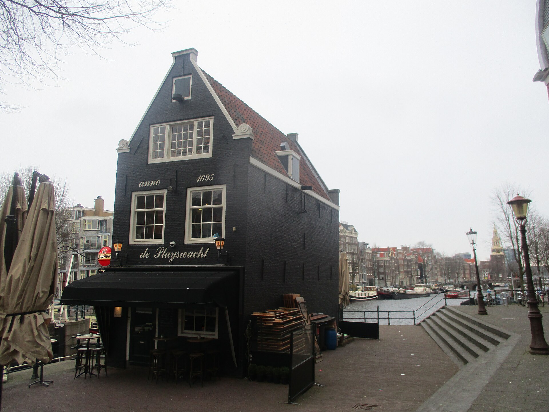 De Sluyswacht stands beside an Amsterdam canal, with closed parasols and stacked outdoor chairs beneath its dark wooden facade.