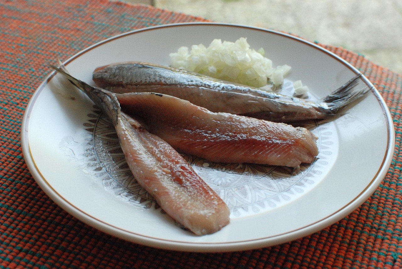 Dutch raw herring fillets with silvery skin and tails lie beside chopped onion on a patterned plate.