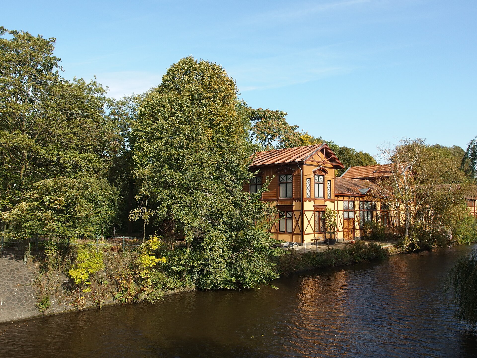 An ochre building with dark timber detailing stands among leafy trees beside the water at ARTIS in Amsterdam.