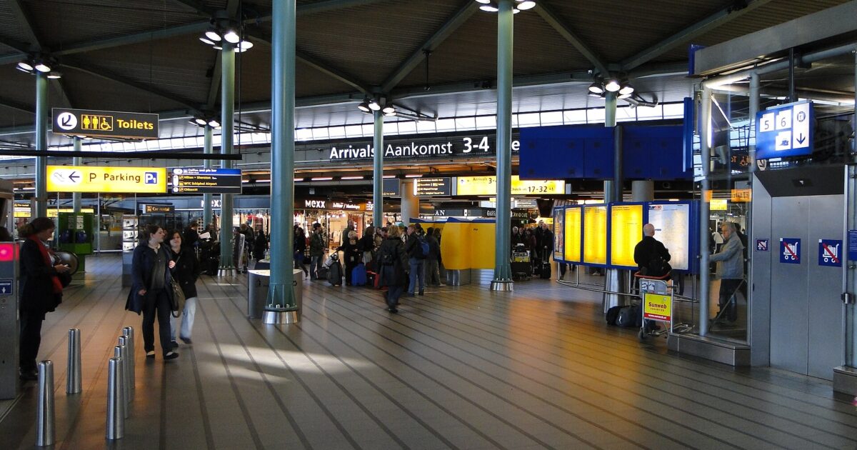 Passengers with luggage gather around illuminated information panels beneath the ribbed roof of Schiphol Plaza at Amsterdam Airport.