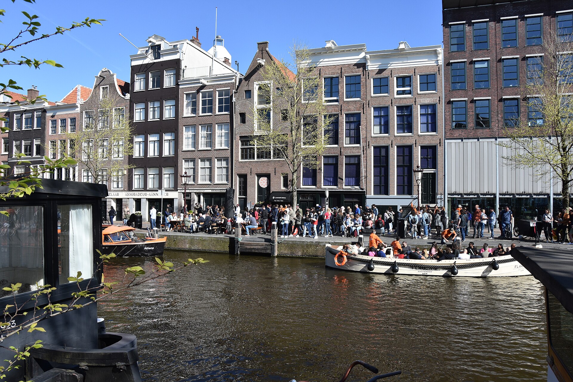 People gather outside the Anne Frank House on Prinsengracht in Amsterdam, while an open passenger boat passes the canal houses.
