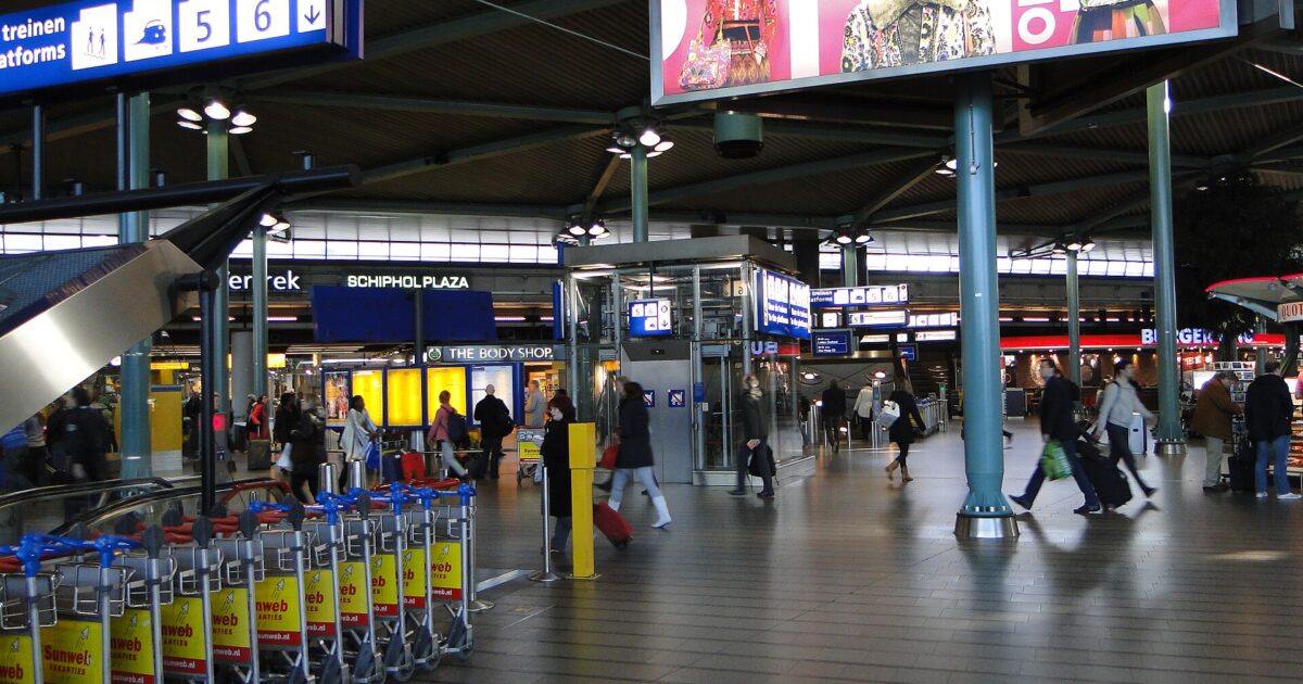 Rows of luggage trolleys stand beside an escalator as passengers cross the tiled concourse at Schiphol Plaza.
