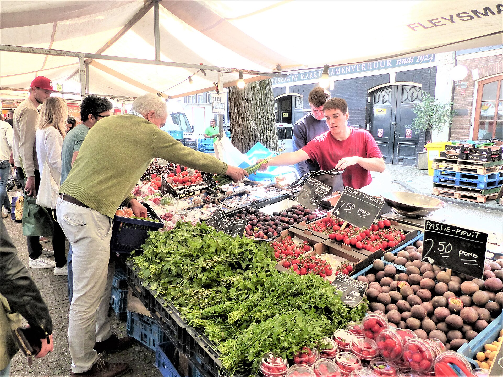A seller serves a customer across a counter of tomatoes, herbs and other produce at Amsterdam Saturday Noordermarkt.