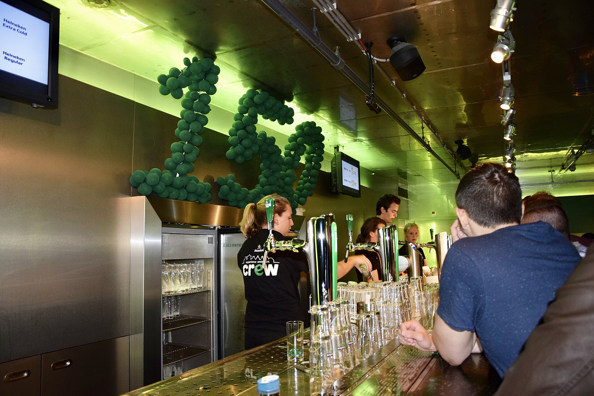 Staff serve visitors at the Heineken Experience bar in Amsterdam, behind rows of glasses and polished beer taps.