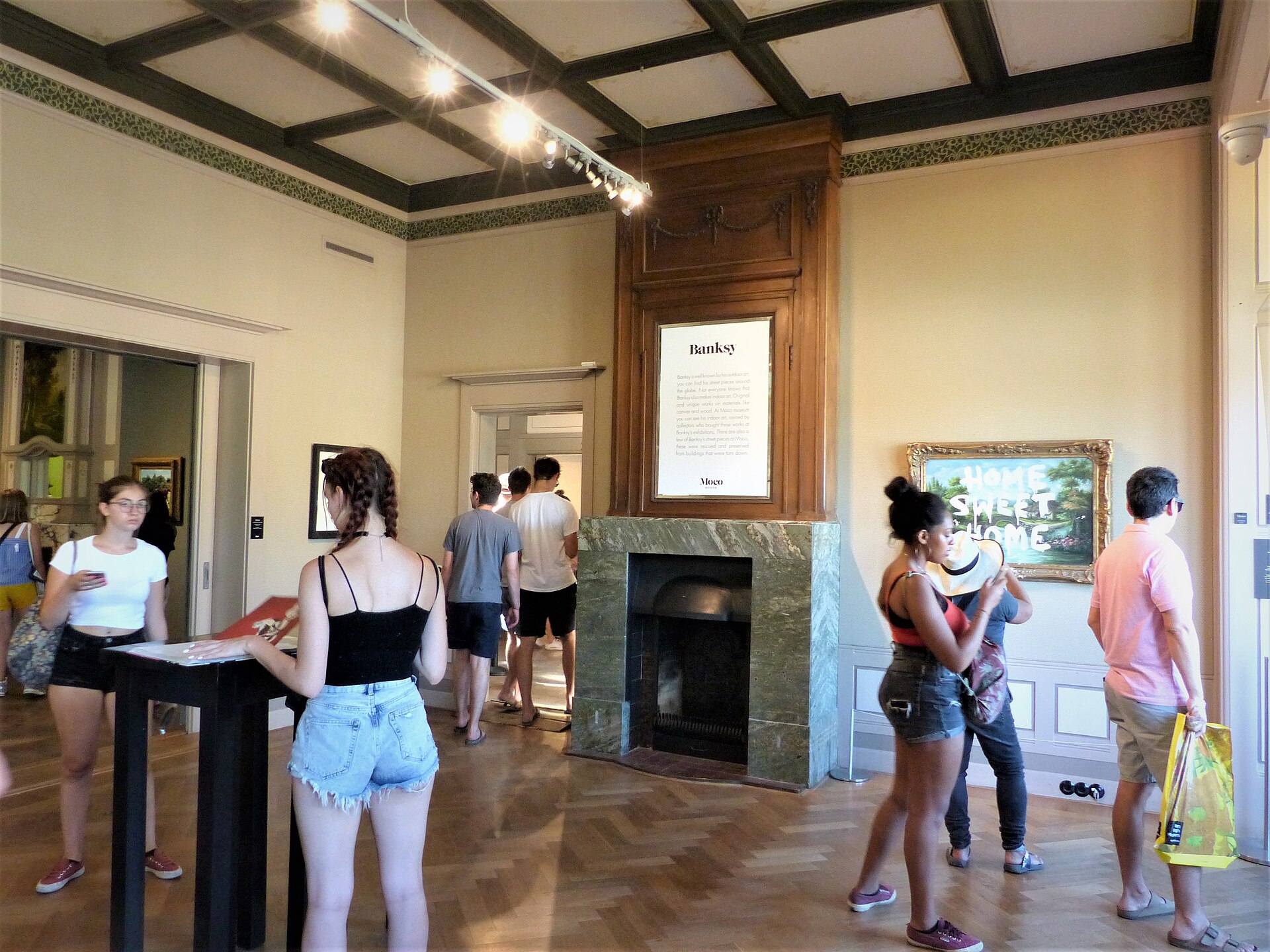 Visitors explore a Moco Museum gallery in Amsterdam, with a parquet floor, coffered ceiling, fireplace and art on the walls.