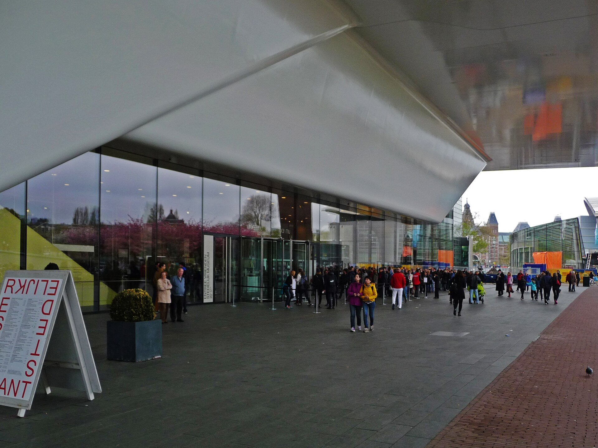 Visitors gather beneath the Stedelijk Museum's projecting white canopy outside its glass entrance on Museumplein in Amsterdam.