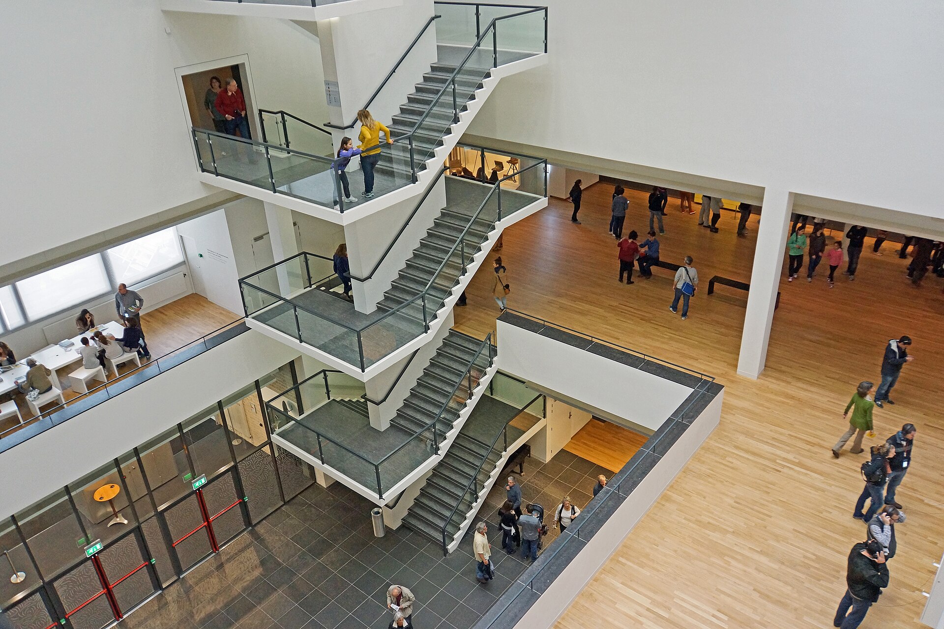 Visitors move between wood-floored galleries and glass-railed staircases inside the Van Gogh Museum in Amsterdam, viewed from above.