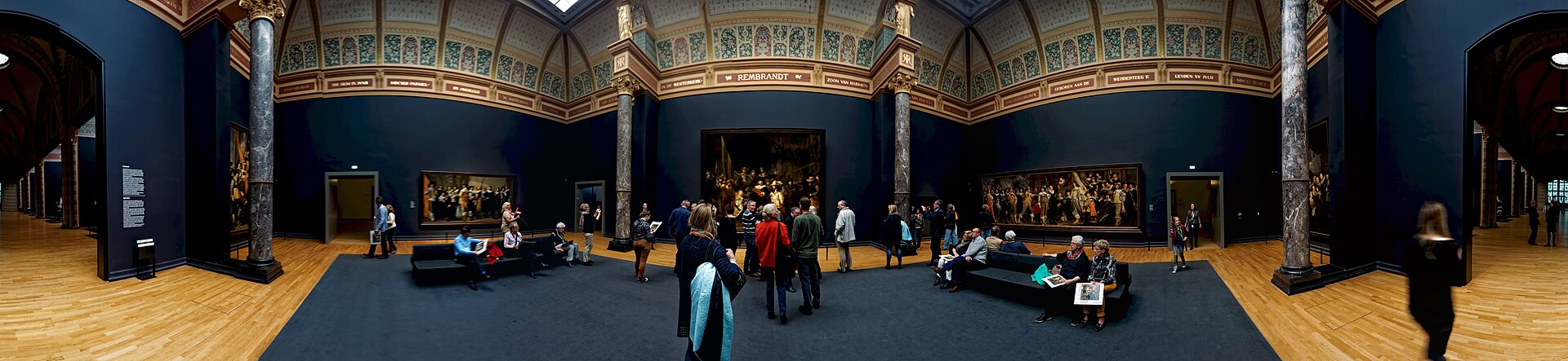 Visitors stand and sit beneath decorated arches in the Rijksmuseum Night Watch Room, with Rembrandt's painting centred on the far wall.