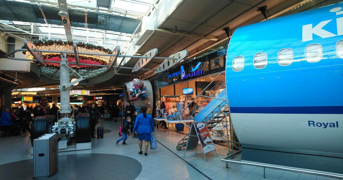 Visitors walk past an aircraft display and souvenir shops beneath the glass roof of Schiphol Plaza.