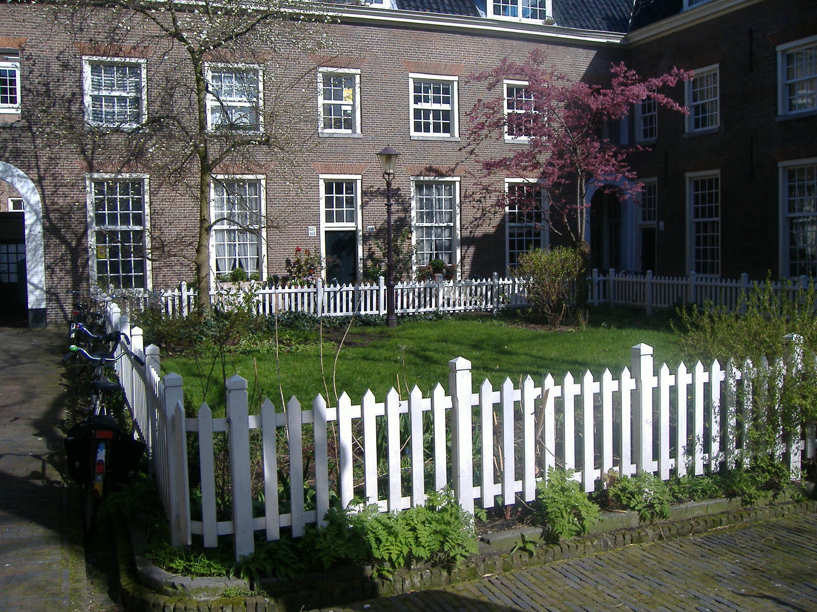 White picket fences frame the garden inside Amsterdam's Karthuizerhof, with tall windows and a flowering tree against brick walls.